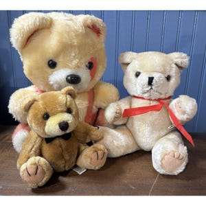 May include: Three plush teddy bears in varying sizes and colors. The largest bear is light tan with a red accent, the middle bear is brown with a black bow tie, and the smallest bear is off-white with a red ribbon.