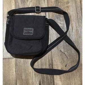 May include: Black crossbody bag with a flap closure and adjustable strap. The bag features a mesh side panel and a small rectangular patch with the brand name "HILFIGER". The strap has white stitching.