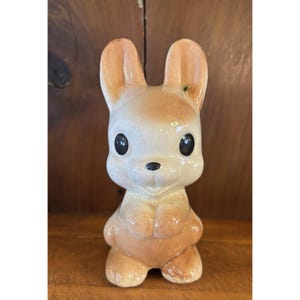 May include: A ceramic figurine of a standing rabbit with a glossy finish. The rabbit is colored in shades of light tan and cream, featuring large, dark, round eyes, a small black nose, and upright ears. The figure has a smooth, rounded body.