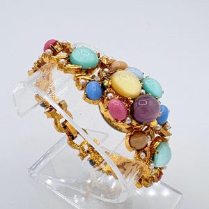 May include: A gold-tone link bracelet featuring a cluster of multicolored cabochon stones in shades of turquoise, pink, purple, yellow, and tan. The design is accented with small white faux pearls set within the textured gold metal framework.