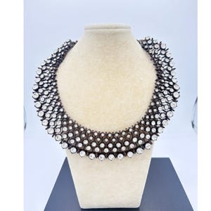 May include: A statement necklace with a dark metal base and numerous clear, round crystals. The necklace is displayed on a cream-colored bust, showcasing its intricate design and sparkling details. The crystals are arranged in a layered pattern.