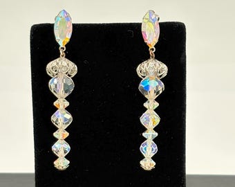 May include: A pair of iridescent crystal earrings. Each earring features an oval-shaped top, a filigree design, and a series of clear, faceted crystals in various shapes. The crystals reflect a rainbow of colors, creating a shimmering effect.