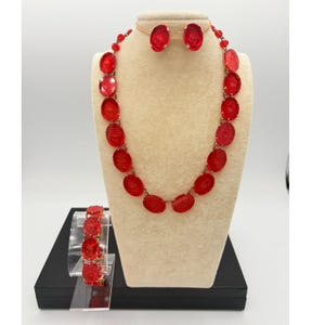 May include: A red jewelry set featuring a necklace, earrings, and bracelet. The necklace and bracelet have oval-shaped red stones. The earrings are also oval-shaped. The jewelry is displayed on a mannequin and a stand against a white background.