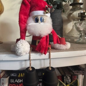May include: A whimsical Santa Claus decoration. The figure features a red plush hat and scarf, a white beard, and blue eyes. Black bell-shaped feet dangle from jute string. The Santa is made from a painted terracotta pot.