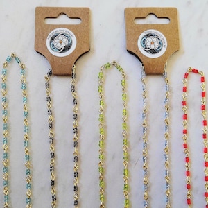 Könnte beinhalten: Mehrere Halsketten mit Perlen in verschiedenen Farben, darunter Blau, Schwarz, Grün und Rot, hängen an braunen Pappanhängern. Die Halsketten haben goldfarbene Ketten und sind mit kleinen, farbigen Perlen verziert. Jedes Etikett hat einen runden Aufkleber mit einem floralen Design und dem Text "FORGET-ME-NOT JEWELRY".