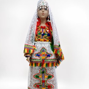 Puede incluir: Maniquí con un vestido de novia marroquí tradicional. El vestido blanco presenta bordados y flecos coloridos. El conjunto incluye un tocado, velo y zapatos a juego. El atuendo se exhibe sobre un fondo blanco.