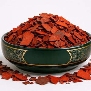May include: A dark green ceramic bowl with gold floral accents filled with a large mound of irregular, flat, reddish-orange flakes. The bowl is rounded with a smooth finish, and several of the same reddish-orange flakes are scattered around the base.