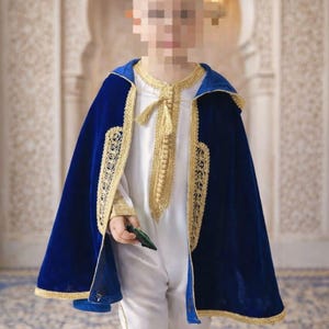 May include: A child's traditional outfit featuring a white tunic with gold embroidery and a royal blue velvet cape. The cape is trimmed with gold and has a hood. The outfit is likely for a special occasion.