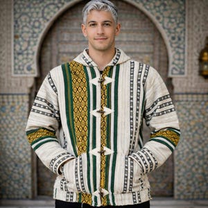 Op de afbeelding: Een crème kleurige jas met capuchon, groene en gouden geometrische patronen en verticale strepen. De jas heeft houten knevelsluitingen en zakken. De mouwen en capuchon hebben gedetailleerde patronen. De jas is gemaakt van een textuurstof.