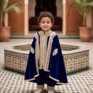 May include: A young child wearing a royal blue velvet outfit with gold embroidery. The outfit includes a cape, jacket, and pants. The child is also wearing white shoes. The background features an ornate courtyard with a fountain.