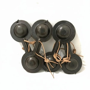 May include: A set of six dark brown castanets, a traditional percussion instrument. Each castanet has a circular shape with a handle and is connected to its pair with a tan leather cord. The instruments are arranged in a group against a white background.