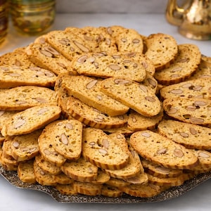 May include: A large platter of golden-brown biscotti cookies, studded with almonds, arranged in a circular pattern. The cookies are on a decorative silver-colored tray, suggesting a dessert or snack. The background includes decorative glass and a marble surface.