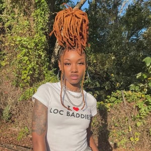 以下が含まれることがあります： 「LOC BADDIES」の文字と赤いハートのグラフィックが入った白いTシャツを着た人。オレンジがかった茶色の長いドレッドヘアを上にスタイリングしています。シルバーのフープイヤリングと複数のシルバーチェーンネックレスを着用しています。