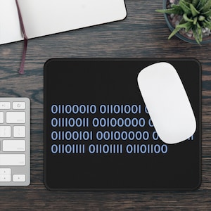 Op de afbeelding: Zwart rechthoekig muismatje met binaire code in lichtblauw. Een witte draadloze muis ligt rechts. Een computertoetsenbord en notitieboek zijn gedeeltelijk zichtbaar. De tekst op de mat luidt: 01100010 01101001, 01110011 00100000, 01100101 00100000, 01101111 01101111 01101100.