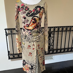 May include: A long-sleeved dress with a beige background and a colorful floral and paisley pattern. The dress has a scoop neckline and a slit up the front, revealing a black and white polka dot panel. The dress is displayed on a white mannequin.