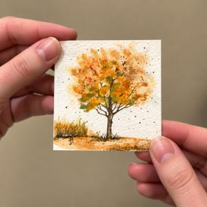 以下が含まれることがあります： 質感のある紙に描かれた小さな正方形の水彩画で、鮮やかなオレンジ、黄色、緑の葉を持つ木が描かれています。木には濃い茶色の幹と枝があり、絵の具が飛び散った明るい背景に配置されています。