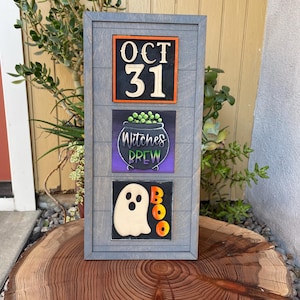 Puede incluir: Un letrero de Halloween de madera gris con tres secciones. La sección superior dice "OCT 31" en blanco sobre un fondo naranja y negro. La sección del medio dice "Witches Brew" encima de un caldero. La sección inferior tiene un fantasma y la palabra "BOO".