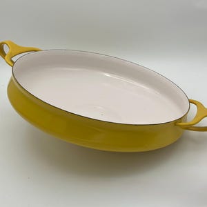 Puede incluir: Una fuente ovalada amarilla con interior blanco y dos asas de bucle. La fuente tiene un acabado brillante y un borde negro. Esta fuente es adecuada para hornear y servir.