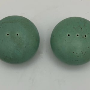 May include: Two round, seafoam green salt and pepper shakers. Each shaker has a speckled surface and a series of small holes on top. The shakers are displayed on a white surface.