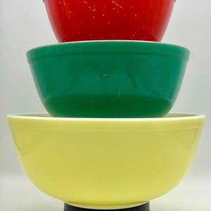 Puede incluir: Una pila de tres cuencos de mezcla vintage en colores primarios: rojo, verde y amarillo. Los cuencos están anidados, con el cuenco rojo en la parte superior, seguido de verde y luego amarillo. Los cuencos son de un material brillante.