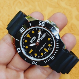 Op de afbeelding: Zwarte en zilveren Seiko automatische duikershorloge met een zwarte rubberen band. De wijzerplaat is zwart met gele markeringen en wijzers. De lunette heeft witte markeringen. Het horloge geeft "DIVERS 200m" en "WED 7" aan.