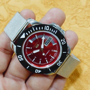 Könnte beinhalten: Eine Seiko Automatik-Uhr aus Edelstahl mit rotem Zifferblatt und schwarzer drehbarer Lünette. Die Uhr hat ein silbernes Mesh-Armband und zeigt den Wochentag an. Das Zifferblatt hat weiße Markierungen und Zeiger.
