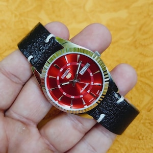 Peut inclure: Une montre-bracelet automatique Seiko 5 dotée d'un cadran rouge éclatant avec des index et des aiguilles argentés. La montre présente une lunette argentée cannelée, un guichet jour-date à trois heures et un bracelet en cuir noir texturé.