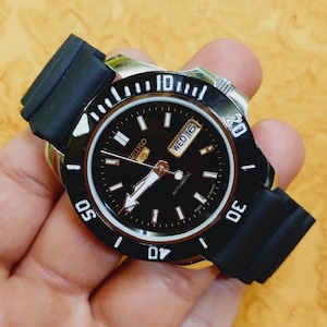 Può includere: Un orologio automatico Seiko nero con cinturino in gomma nero. Il quadrante dell'orologio è nero con indici e lancette bianche. La lunetta ha segni bianchi. Il giorno e la data sono visualizzati alle 3. L'orologio è tenuto in una mano.