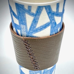 May include: A brown leather coffee cup sleeve with a stitched seam. The sleeve is wrapped around a white paper coffee cup with a blue geometric pattern. The text "CONTAINS NO POLYstyrene FOAM" is visible on the cup.