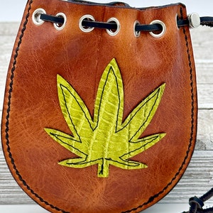 May include: Brown leather pouch with a green cannabis leaf applique. The pouch has a drawstring closure and metal grommets. The edges are stitched with black thread. The pouch is approximately 5 inches tall and 4 inches wide.