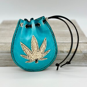May include: A turquoise leather pouch with a black drawstring closure. The pouch features a beige cannabis leaf design and black stitching around the edges. The pouch is approximately 4 inches tall.