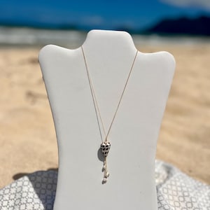 Puede incluir: Un collar de cadena dorada con un colgante con una concha marina manchada en blanco y negro. El collar tiene un diseño colgante con pequeñas perlas blancas. El collar se exhibe en un maniquí blanco sobre un fondo de playa.