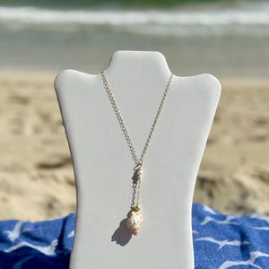 May include: A silver necklace with a seashell pendant displayed on a white bust. The necklace has a delicate chain with a small pearl and a larger seashell pendant. The background shows a beach with sand and ocean waves.