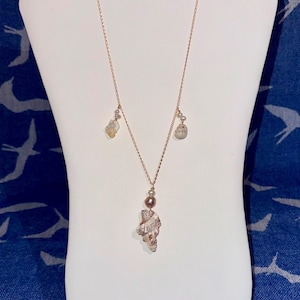May include: A delicate necklace with a gold chain, featuring a seashell pendant, a pearl, and small, irregular-shaped beads. The necklace is displayed on a white mannequin against a blue background.