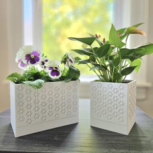 May include: Two white rectangular and square planters with a floral pattern. The rectangular planter holds pansies with purple and white petals. The square planter contains a green plant with red accents. Both planters sit on a dark surface.