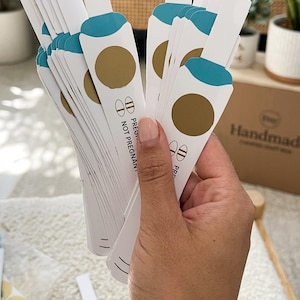 Può includere: Una collezione di repliche di test di gravidanza in carta bianca con una sezione superiore color ottanio, un'area circolare dorata da grattare e testo stampato che recita PREGNANT e NOT PREGNANT con simboli. Le carte sono tenute a ventaglio.