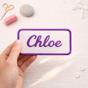 May include: A rectangular white patch with a purple border and the name "Chloe" embroidered in purple script. The patch is held in a hand, with sewing supplies like scissors and thread in the background.
