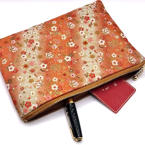 Peut inclure: Une pochette orange à motifs floraux avec une fermeture éclair dorée, un stylo noir et une carte rouge partiellement visible. La pochette porte une étiquette marron avec les lettres "JN". Le motif floral comprend des fleurs blanches, rouges et dorées.