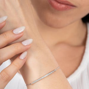Peut inclure: Un bracelet en argent délicat avec un design linéaire, orné de petits pierres précieuses scintillantes. Le bracelet est présenté sur un poignet, mettant en valeur son style élégant et minimaliste. Les pierres précieuses sont disposées en rangée.