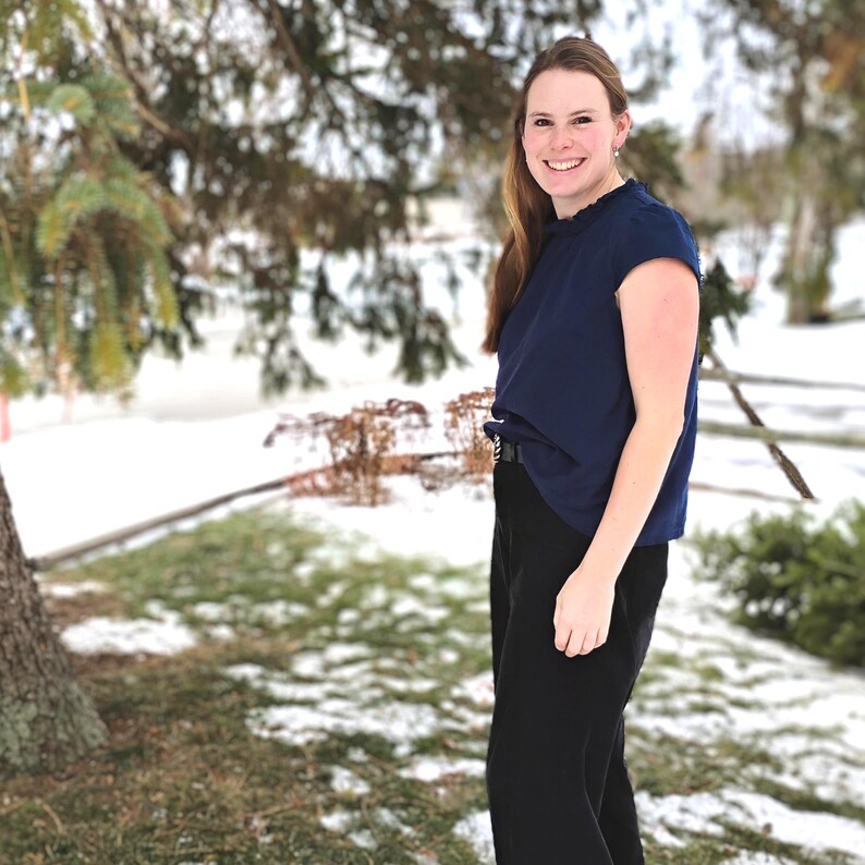 May include: A woman wearing a navy blue top with a ruffled neckline and black pants. The top is tucked into the pants, and a belt is visible. The background shows a snowy outdoor scene with trees.