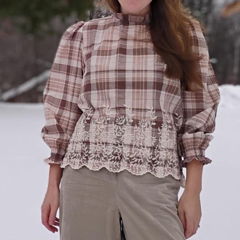 May include: A plaid blouse in shades of brown and cream, featuring a high neckline and long sleeves with ruffled cuffs. The bottom of the blouse has a white floral lace trim. The blouse is paired with light green pants.