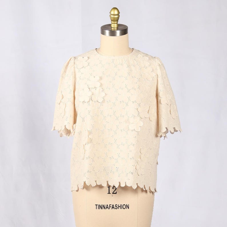 May include: A cream-colored, short-sleeved top with a floral lace design. The top has a scalloped hem and sleeves. The text "12" and "TINNAFASHION" are visible on the mannequin.