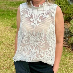 May include: A sleeveless, white lace top with a high, ruffled neckline. The intricate lace design features floral and geometric patterns. The top is made of a delicate, semi-sheer fabric, offering a vintage or bohemian aesthetic.