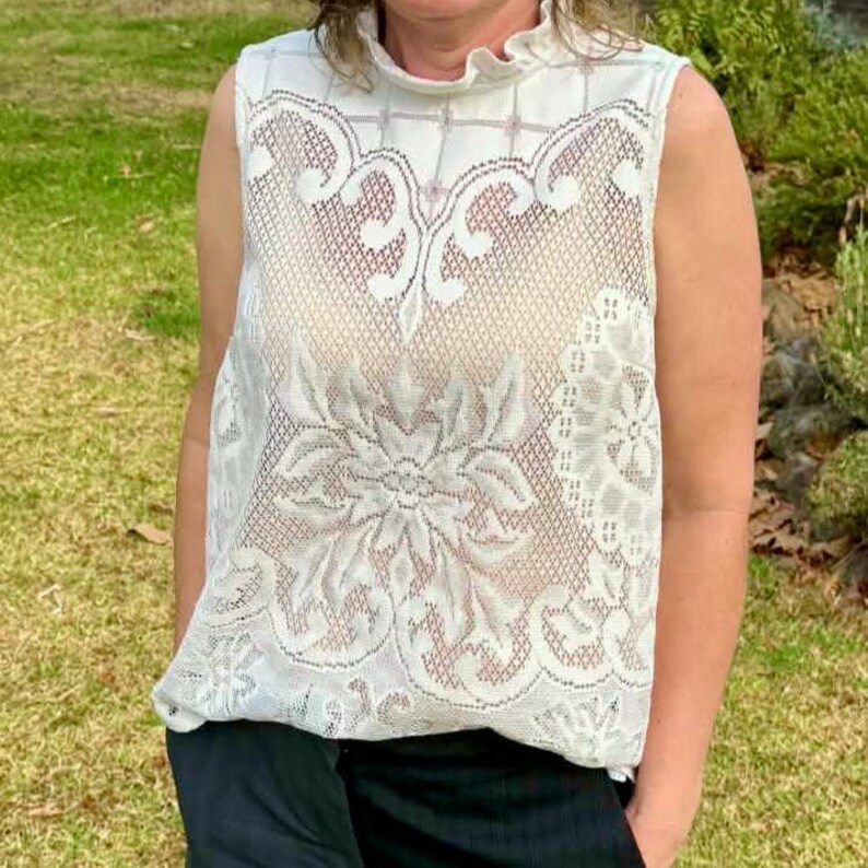May include: A sleeveless, white lace top with a high, ruffled neckline. The intricate lace design features floral and geometric patterns. The top is made of a delicate, semi-sheer fabric, offering a vintage or bohemian aesthetic.