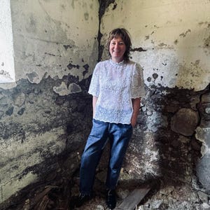 May include: A white lace top with short sleeves and a high neckline is paired with blue jeans. The top features an intricate floral pattern. The person is wearing black shoes. The background is a weathered stone wall.