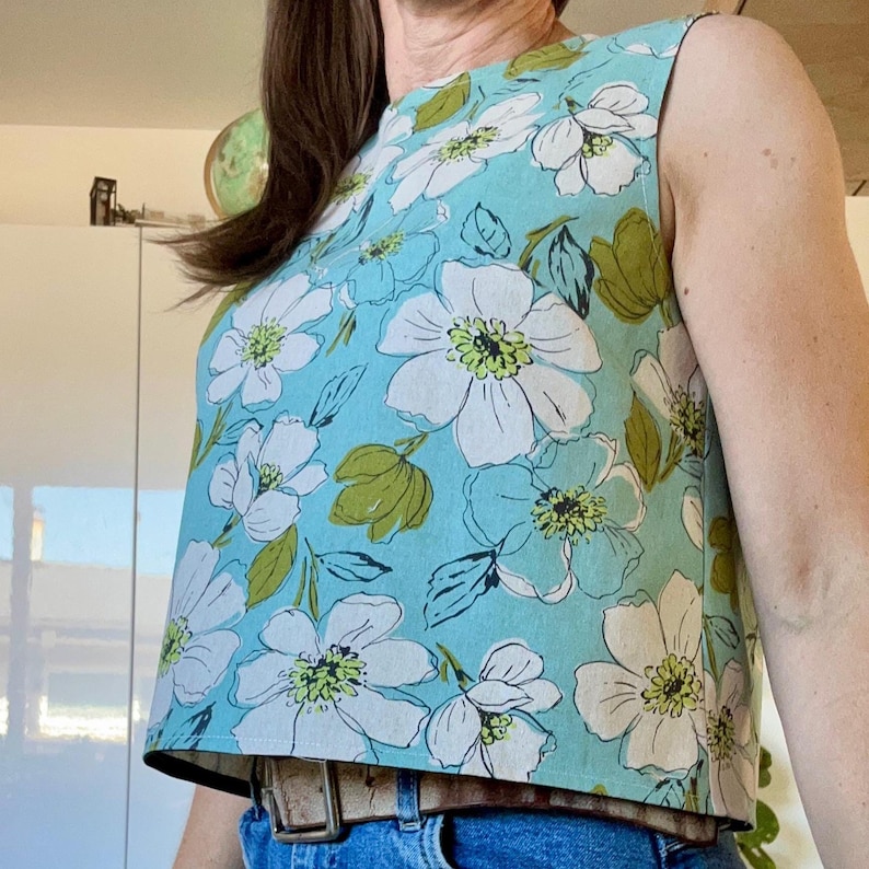 May include: A sleeveless top with a light blue background and a large white floral pattern with green leaves. The top is paired with blue jeans and a brown belt.