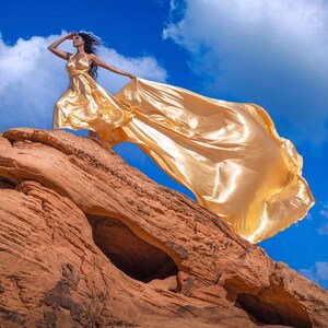 May include: A flowing, golden-yellow satin gown with a halter neckline and a long, trailing skirt. The dress is worn on a red rock formation against a bright blue sky with fluffy white clouds. The fabric billows in the wind.