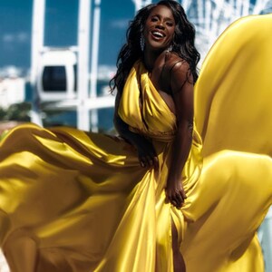 May include: A woman in a flowing, golden-yellow dress smiles against a backdrop of a Ferris wheel and a bright blue sky. The dress has a halter neckline and a high slit, creating a dramatic effect. The fabric appears to be satin or silk.