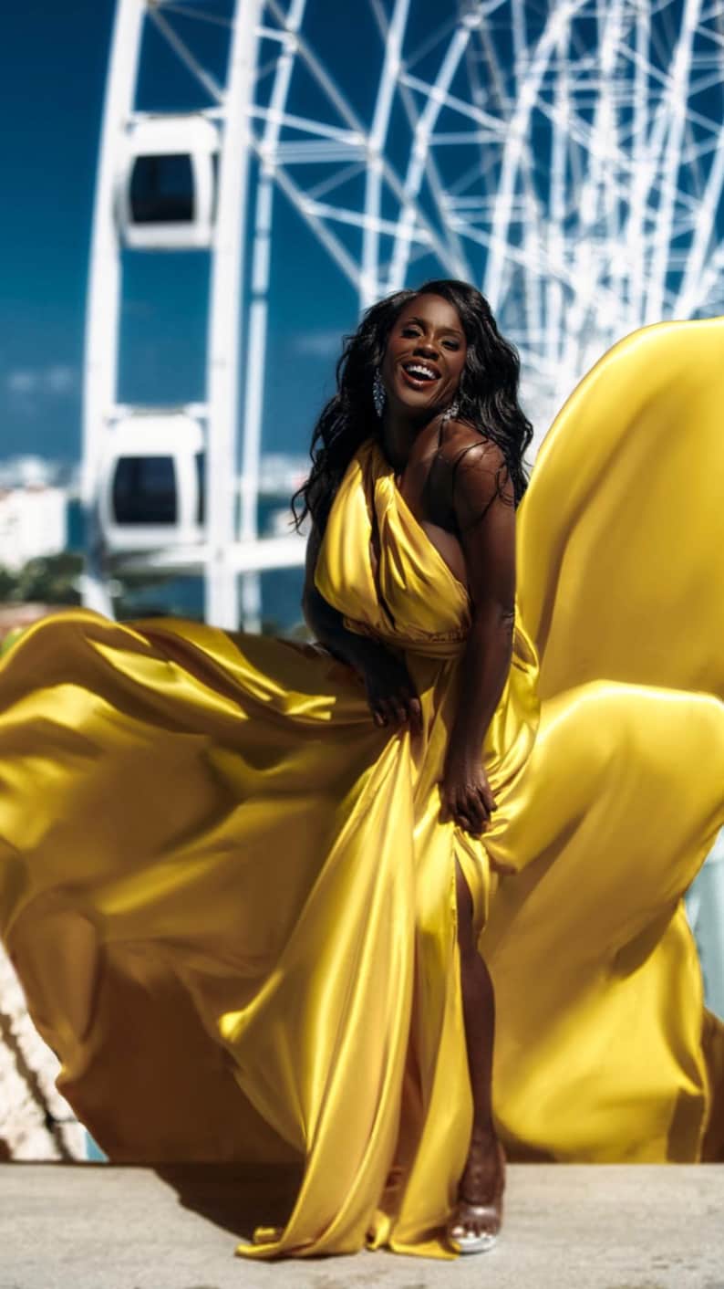 May include: A woman in a flowing, golden-yellow dress smiles against a backdrop of a Ferris wheel and a bright blue sky. The dress has a halter neckline and a high slit, creating a dramatic effect. The fabric appears to be satin or silk.
