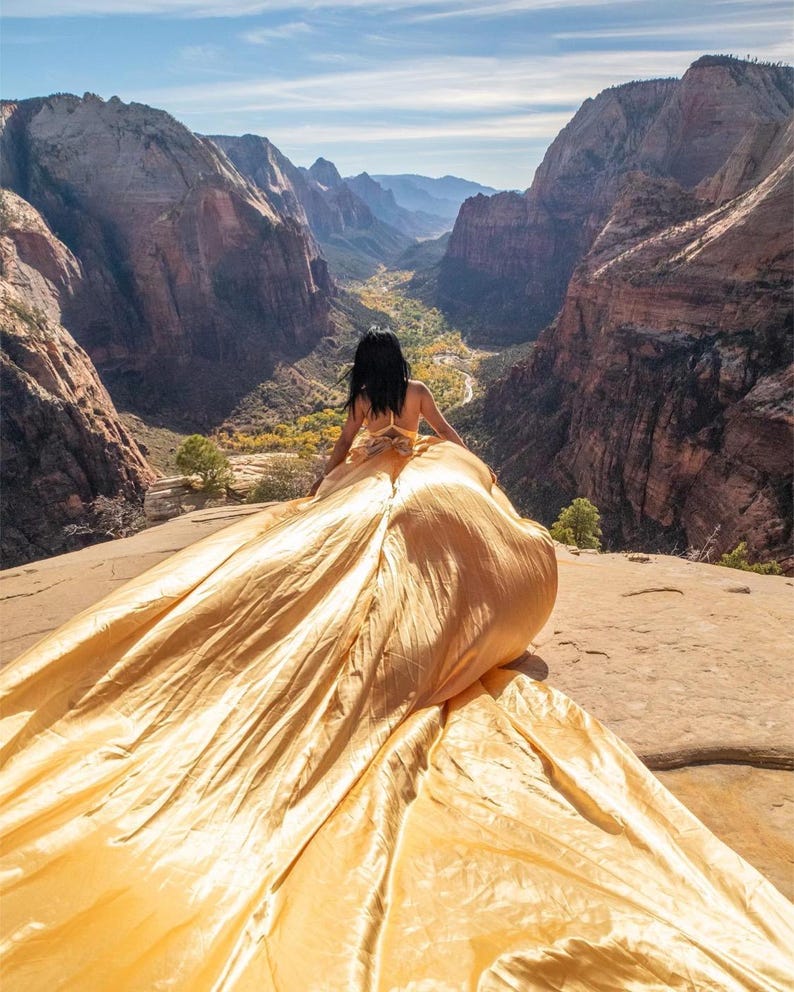 May include: A woman in a golden gown sits on a rock overlooking a canyon. The flowing fabric of the dress contrasts with the rugged landscape of the mountains and valley below. The sky is blue with a few clouds.